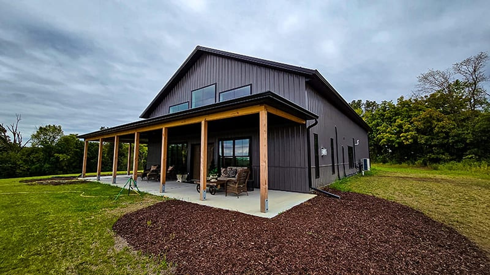 The Cedar barndominium with dark board-and-batten steel siding, a black metal roof, and a covered wraparound porch